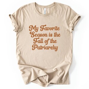 May include: Beige t-shirt with rolled sleeves. The shirt features the text "My Favorite Season is the Fall of the Patriarchy" in a brown, retro-style font. The shirt is made of a soft material and is suitable for casual wear.