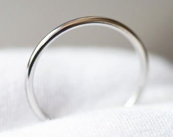 Può includere: Primo piano di un anello sottile d'argento. L'anello è liscio e lucido, con una fascia arrotondata. Poggia su un tessuto bianco e strutturato, creando un contrasto. Il design dell'anello è minimalista ed elegante, adatto all'uso quotidiano.