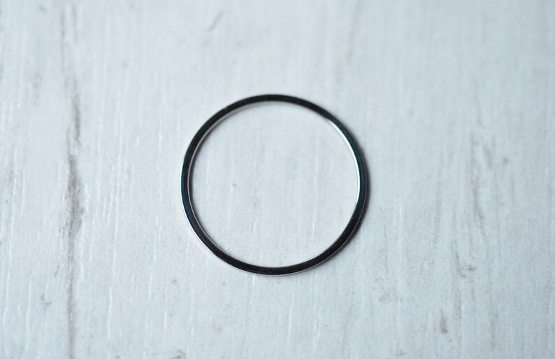 Pu&ograve; includere: Un anello di metallo color argento, posto piatto su una superficie di legno bianco.