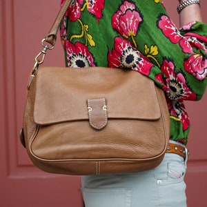 May include: A brown leather shoulder bag with a flap closure and a gold buckle. The bag is worn by a person wearing a green and pink floral blouse and light green jeans.