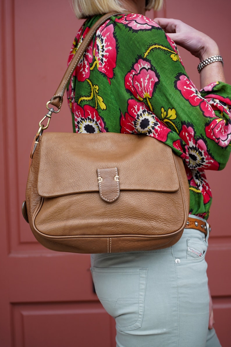 May include: A brown leather shoulder bag with a flap closure and a gold buckle. The bag is worn by a person wearing a green and pink floral blouse and light green jeans.