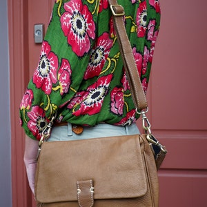 May include: A brown leather shoulder bag with a flap closure and a buckle. The bag has a long strap and is worn over the shoulder of a person wearing a green and red floral blouse.