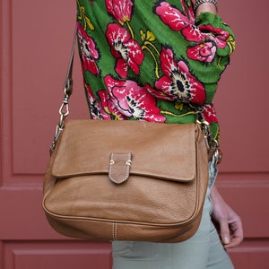 May include: A brown leather shoulder bag with a flap closure and a gold buckle. The bag is worn by a person wearing a green and red floral blouse and light green pants.