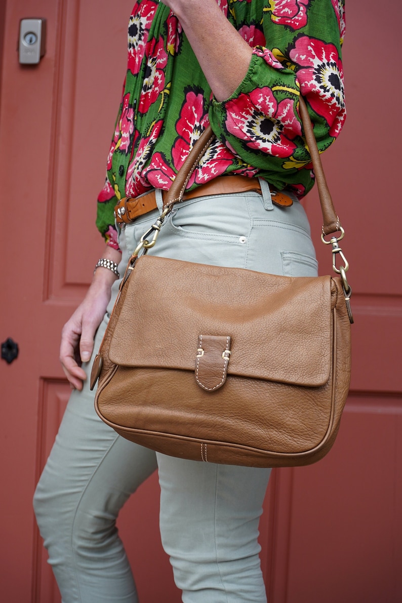 May include: A woman wearing a green and red floral blouse with a brown leather shoulder bag. The bag has a flap closure and a long strap.
