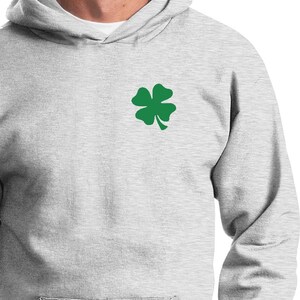 Puede incluir: Una sudadera con capucha gris con un trébol de cuatro hojas verde bordado en el pecho izquierdo.