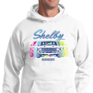 May include: White hooded sweatshirt featuring a colorful graphic of a Shelby car with the word "Shelby" in a stylized font. The design includes the text "Shelby" and "Shelby" in a rectangular box. A classic car-themed apparel item.