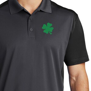 Puede incluir: Una camiseta polo negra y gris con un trébol de cuatro hojas verde bordado en el pecho izquierdo.