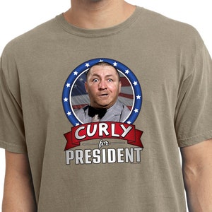 May include: A khaki t-shirt with a graphic of Curly from The Three Stooges. The graphic features Curly's face in a circle with red, white, and blue stars. The text "Curly for President" is below the graphic.