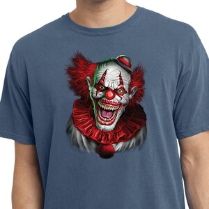 May include: A blue t-shirt featuring a scary clown face graphic. The clown has red hair, a red nose, and a wide, toothy grin.