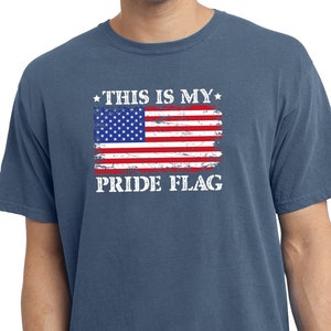 Puede incluir: Una camiseta azul con un gráfico de bandera estadounidense desgastado y el texto "*THIS IS MY* PRIDE FLAG" impreso en la parte delantera.