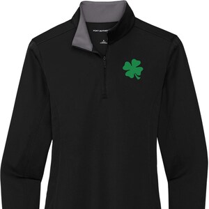 Puede incluir: Camiseta negra de manga larga con un diseño de trébol de cuatro hojas verde. La camiseta tiene un cuello con cremallera y está hecha de tejido de rendimiento.