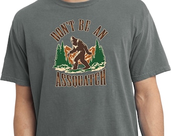 Może przedstawiać: Szara koszulka z grafiką Bigfoota idącego przed górskim krajobrazem. Tekst "DON'T BE AN ASSQUATCH" jest wydrukowany nad i pod obrazem w brązowej, stylizowanej czcionce.