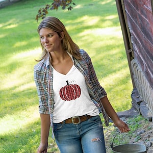 May include: A woman wearing a white t-shirt with a red and black plaid pumpkin graphic, blue jeans, and green rain boots. She is carrying two metal buckets.