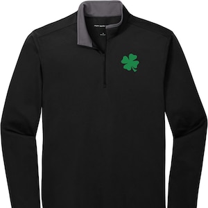 Puede incluir: Camiseta negra de manga larga con un diseño de trébol de cuatro hojas verde. La camisa tiene un cuello con cremallera y un interior gris.