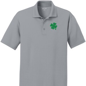 Puede incluir: Una camisa polo de manga corta gris con un trébol de cuatro hojas verde bordado en el pecho izquierdo.