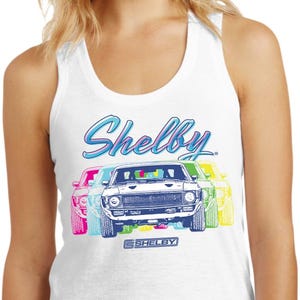 May include: White tank top featuring a colorful graphic of three Shelby Mustang cars. The word "Shelby" is written in a blue script font above the cars, with the Shelby logo below. The cars are outlined in pink, blue, and yellow.