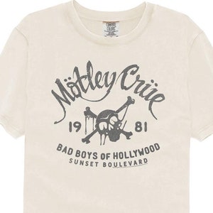 May include: A white t-shirt with a black and gray graphic print of a skull and crossbones with the text "Motley Crue 1981 Bad Boys of Hollywood Sunset Boulevard".