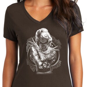 May include: A brown V-neck t-shirt with a graphic print of a woman with tattoos and a bandana covering her face. The woman is surrounded by dollar bills.