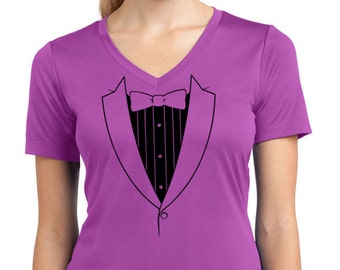 Puede incluir: Una camiseta morada de cuello en V con un gráfico de esmoquin negro en la parte delantera. El gráfico incluye una pajarita, una solapa de chaqueta y un botón.