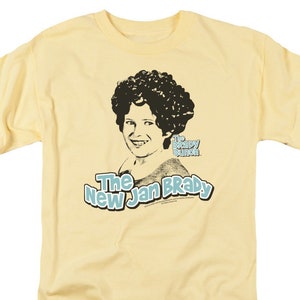 Pu&ograve; includere: Una maglietta gialla con un'immagine in bianco e nero di una donna con un'ampia acconciatura afro. Il testo "The New Jan Brady" &egrave; stampato in blu sulla maglietta. Il testo "The Brady Bunch" &egrave; stampato in nero sulla maglietta.