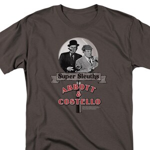 May include: A dark gray t-shirt with a graphic of Abbott and Costello in a magnifying glass. The text "Super Sleuths" is above the image and "Abbott & Costello" is below the image.
