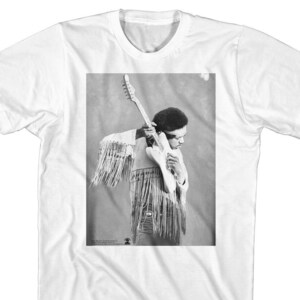 May include: Black and white photograph of a musician playing a guitar. The musician is wearing a fringed jacket and has long hair. The photograph is printed on a white t-shirt.