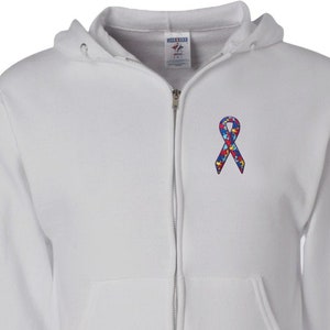 May include: White zip-up hoodie with a hood and front pockets. A colorful ribbon emblem, featuring puzzle piece designs in red, blue, yellow, and green, is embroidered on the left chest. The hoodie has a full zipper closure and ribbed cuffs.