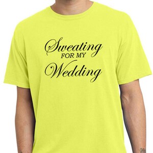 May include: Neon yellow t-shirt with the text "Sweating for my Wedding" in black script. The shirt is a crew neck style and is made of a soft material. The bottom of the shirt is visible.