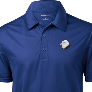 May include: A royal blue short-sleeved polo shirt with a white embroidered bald eagle on the left chest. The shirt has a button-down collar and a Sport-Tek brand label.