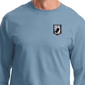 Può includere: Una t-shirt blu stonewashed a maniche lunghe con una piccola toppa ricamata sul petto sinistro. La toppa è in bianco e nero e presenta una silhouette di un soldato con le parole "POW-MIA You Are Not Forgotten" stampate su di essa.