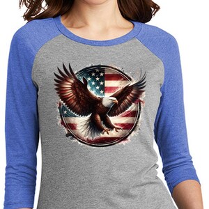 May include: A gray raglan shirt with blue sleeves featuring a detailed graphic of an American bald eagle with outstretched wings, set against a backdrop of the American flag. The shirt is a three-quarter sleeve style.