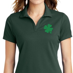 Puede incluir: Polo de color verde oscuro con un emblema de trébol verde en el pecho izquierdo. La camisa tiene cuello y mangas cortas. La modelo lleva vaqueros azul oscuro.