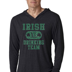May include: Dark gray long-sleeve hooded shirt with the text "IRISH DRINKING TEAM" in distressed green lettering. The design includes a green oval with shamrocks. The shirt has a V-neck and a relaxed fit.