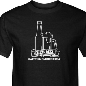Peut inclure: T-shirt noir avec un contour blanc d'une bouteille de bière et d'une chope à bière. Le texte "Beer Me! Happy St. Patrick's Day" est imprimé sur le t-shirt.