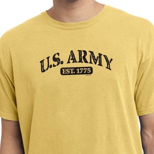 Puede incluir: Una camiseta amarilla con un gráfico negro desgastado que dice "U.S. ARMY EST. 1775".