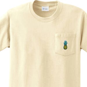 May include: A natural colored t-shirt with a single pocket on the left side. The pocket has a small embroidered pineapple design.