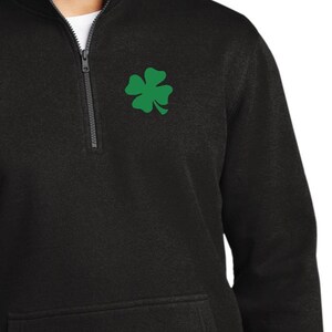 Puede incluir: Sudadera negra con cremallera de cuarto de cuello y un emblema de trébol verde en el pecho izquierdo. La sudadera tiene un bolsillo delantero y una cremallera que llega hasta el cuello. El modelo lleva vaqueros azules.