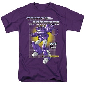 May include: Purple t-shirt with a graphic of Shockwave, a Decepticon from the Transformers cartoon series. The graphic shows Shockwave holding a gun and standing on a grid pattern. The text "The Transformers" and "Decepticon Shockwave" are also on the shirt.