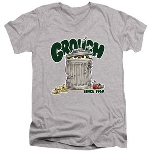 May include: A gray V-neck t-shirt with a green cartoon of Oscar the Grouch peeking out of a trash can. The text "Grouch" is above the trash can and "Since 1969" is below the trash can.