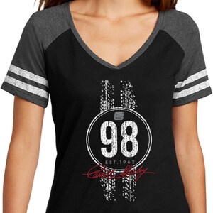 May include: Black V-neck t-shirt with grey raglan sleeves and white stripes. The shirt features a white graphic with the number 98 inside a circle, tyre track designs, and the text "Est. 1962".