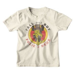 May include: A white t-shirt with a vintage-style graphic of two bears in a red and yellow circle. The text "Fall Out Boy" is written around the circle, and the text "Folie a Deux" is written below the circle.