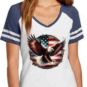May include: A white, navy blue, and gray women's t-shirt with a baseball-style collar. The shirt features a graphic of a bald eagle flying in front of an American flag.