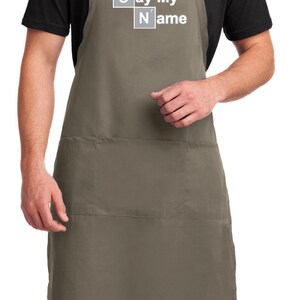 May include: A khaki-colored apron with a large pocket on the front. The apron has a white graphic that says "Say My Name" with the letters S and N styled as chemical element boxes.