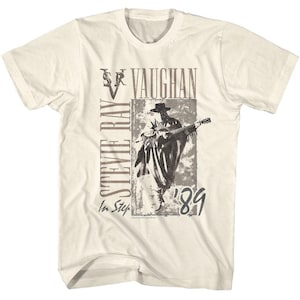 May include: Cream-colored t-shirt featuring a sepia-toned image of a musician playing a guitar, with the text "STEVIE RAY VAUGHAN" and "In Step '89" in a distressed font. The shirt has short sleeves and a crew neck.