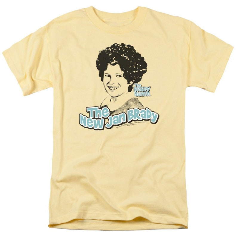 Pu&ograve; includere: Una maglietta gialla con un'immagine in bianco e nero di una donna con un'ampia acconciatura afro. Il testo "The New Jan Brady" &egrave; stampato in blu sulla maglietta. Il testo "The Brady Bunch" &egrave; stampato in nero sulla maglietta.