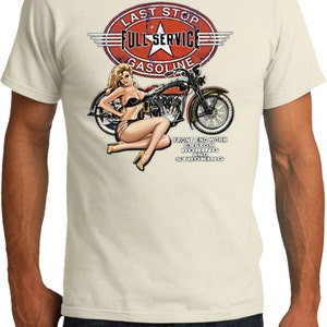 Op de afbeelding: Een wit T-shirt met een vintage-stijl illustratie van een pin-up meisje dat op een motorfiets zit. De illustratie bevat een rood, wit en blauw tankstationbord met de tekst "Last Stop Full Service Gasoline". De tekst onder de illustratie luidt "Front End Work Custom Boring and Stroking".