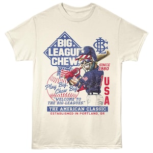 May include: Cream-colored t-shirt featuring a baseball player graphic. The design includes the text "BIG LEAGUE CHEW" and "USA" with a baseball and the words "Play Big, Chew Big". The shirt is a classic American design.