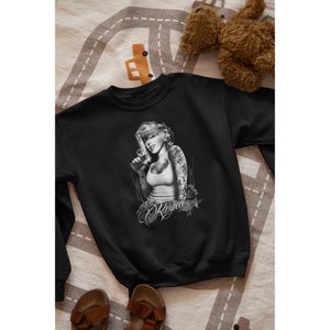 Puede incluir: Sudadera negra con un estampado gráfico de una mujer con los ojos vendados y una pistola, con la palabra "Respect" escrita debajo de ella.