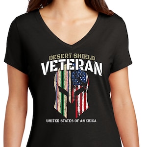 Puede incluir: Camiseta negra de cuello en V con un gráfico desgastado de un casco espartano con la bandera estadounidense y una cinta militar. El texto "Desert Shield Veteran" y "United States of America" está impreso en la camiseta.