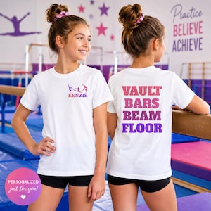 May include: A white short-sleeved t-shirt featuring a small graphic of gymnasts and the name KENZIE on the front left chest. The back of the shirt displays the words VAULT, BARS, BEAM, and FLOOR in a vertical list with a pink to purple gradient.
